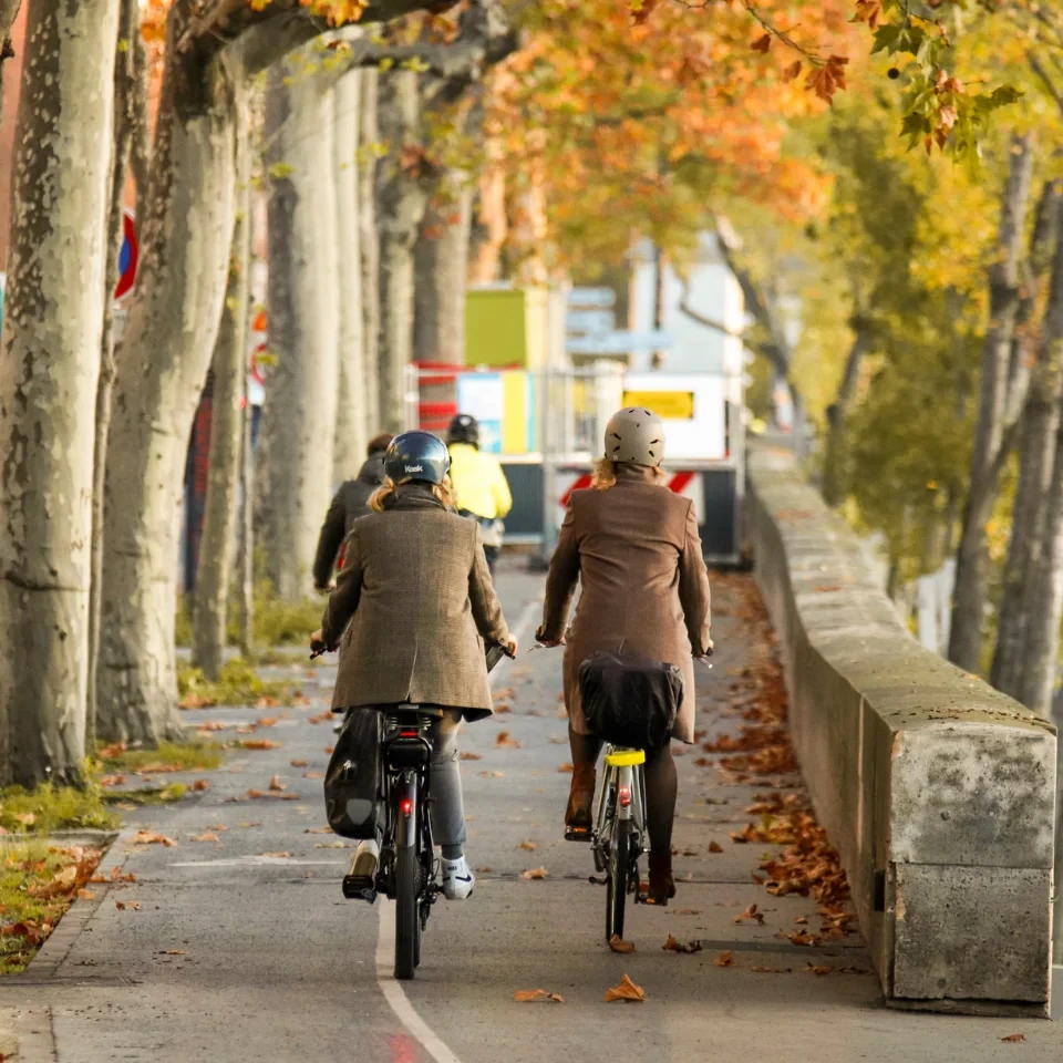 Deux cyclistes casqués roulant sur une piste cyclable urbaine bordée d’arbres en automne